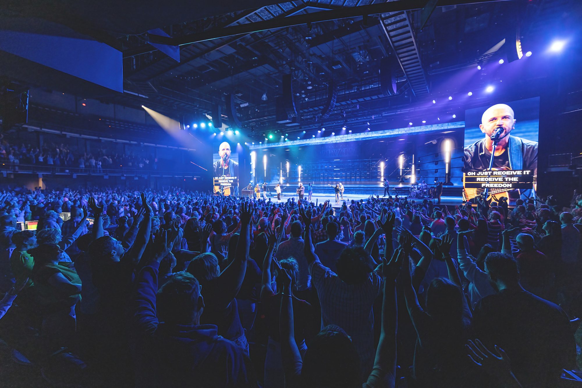 A packed auditorium at Crossroads Church with hands raised during worship, the stage washed in blue light and the worship leader carried to two large side screens.